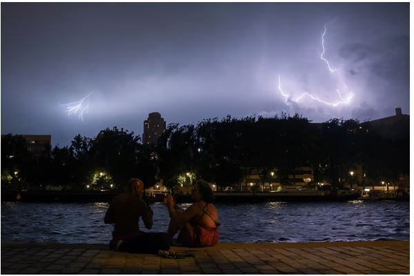 People sit on the banks of the Bassin de la Villette during a storm, as lightning strikes behind them amid a heatwave in Paris