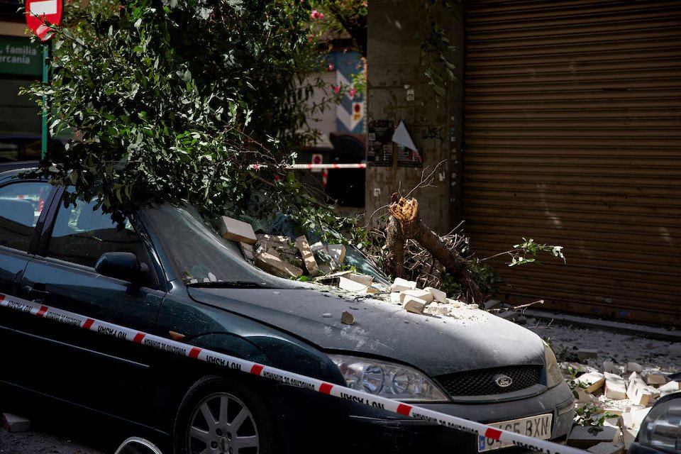 A fallen tree branch lies atop a damaged vehicle on a street